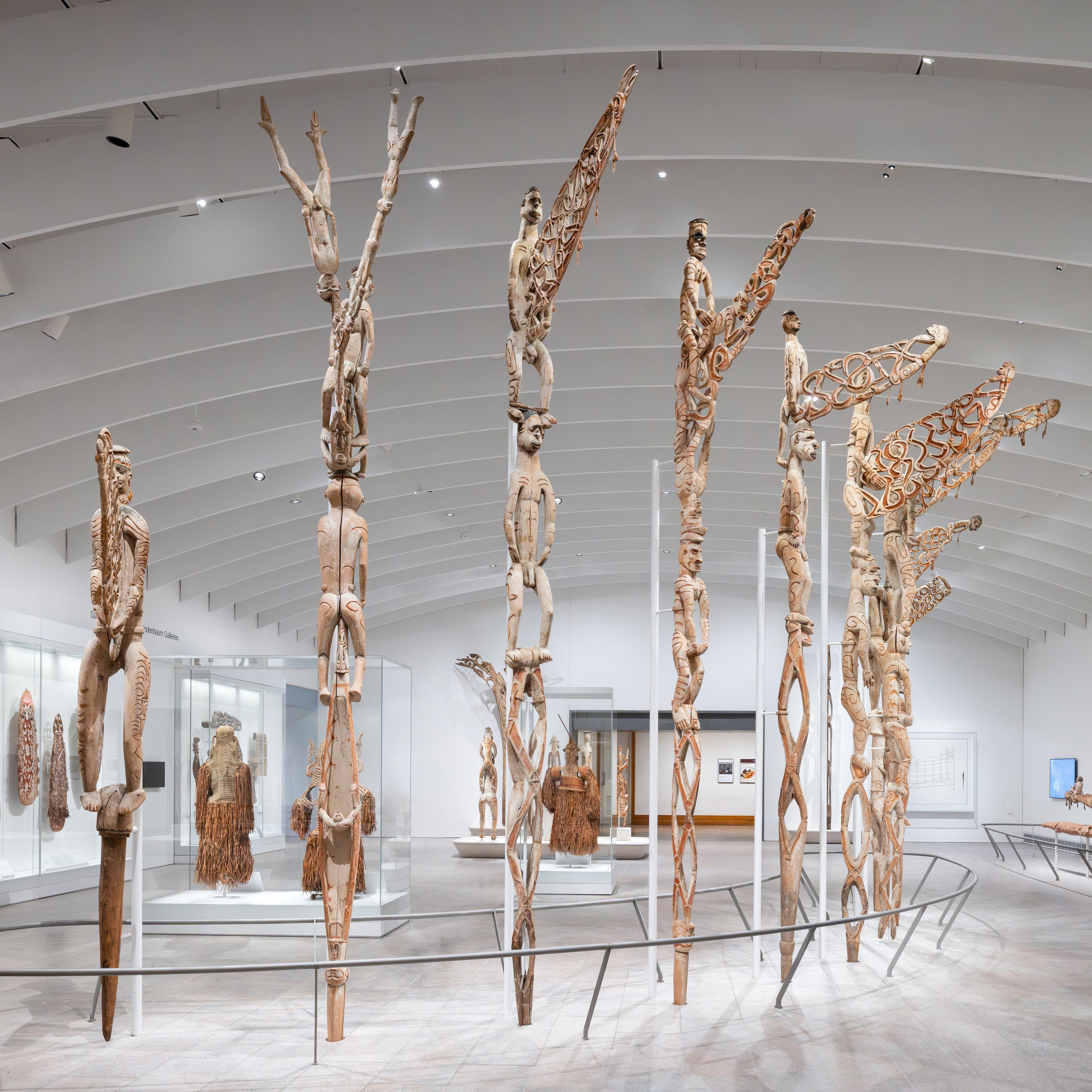 Tall ancestor poles arranged in a curve in the center of a bright white gallery space.
