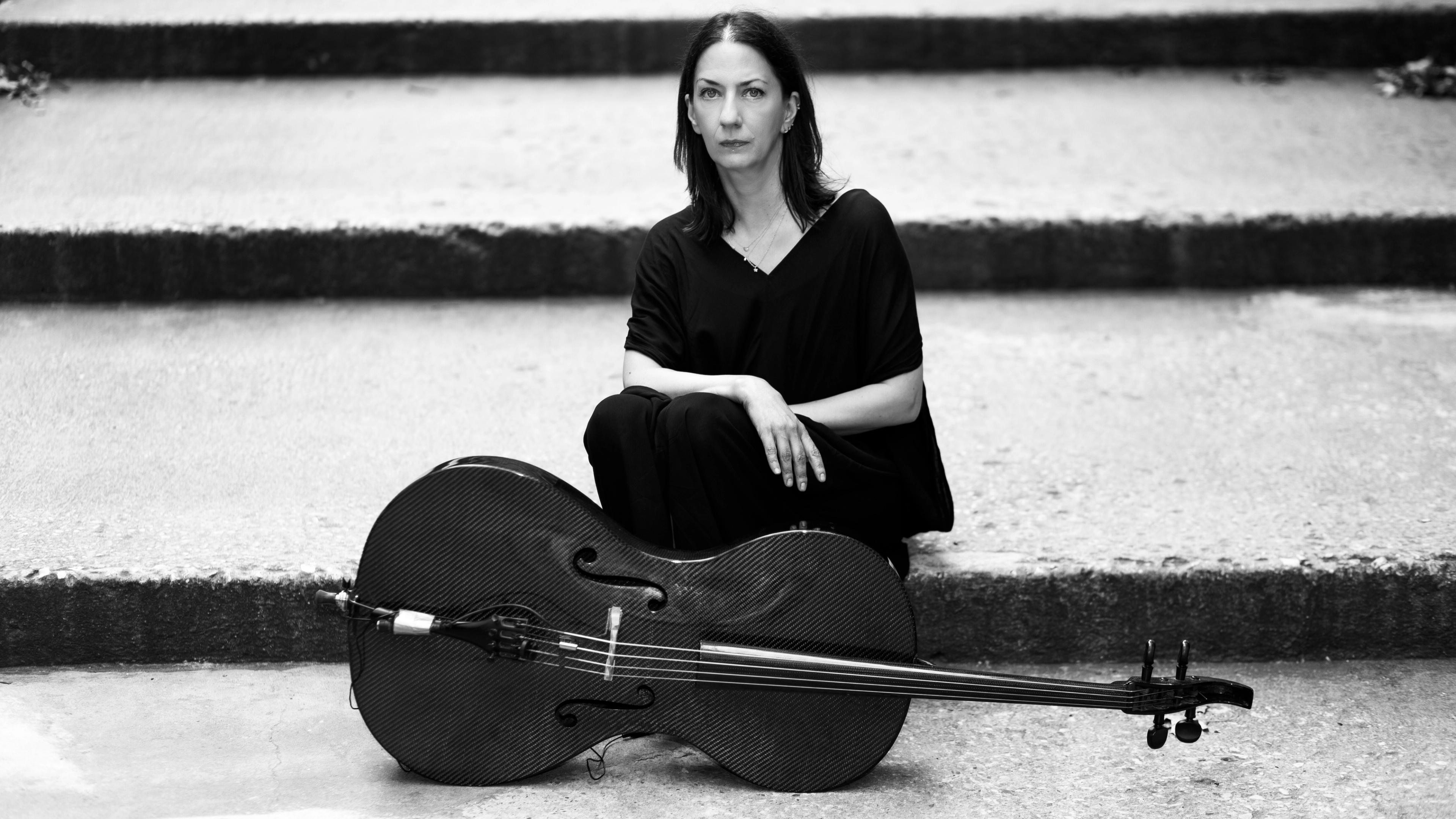 A black-and-white image of a woman sitting next to a carbon fiber cello on its side.