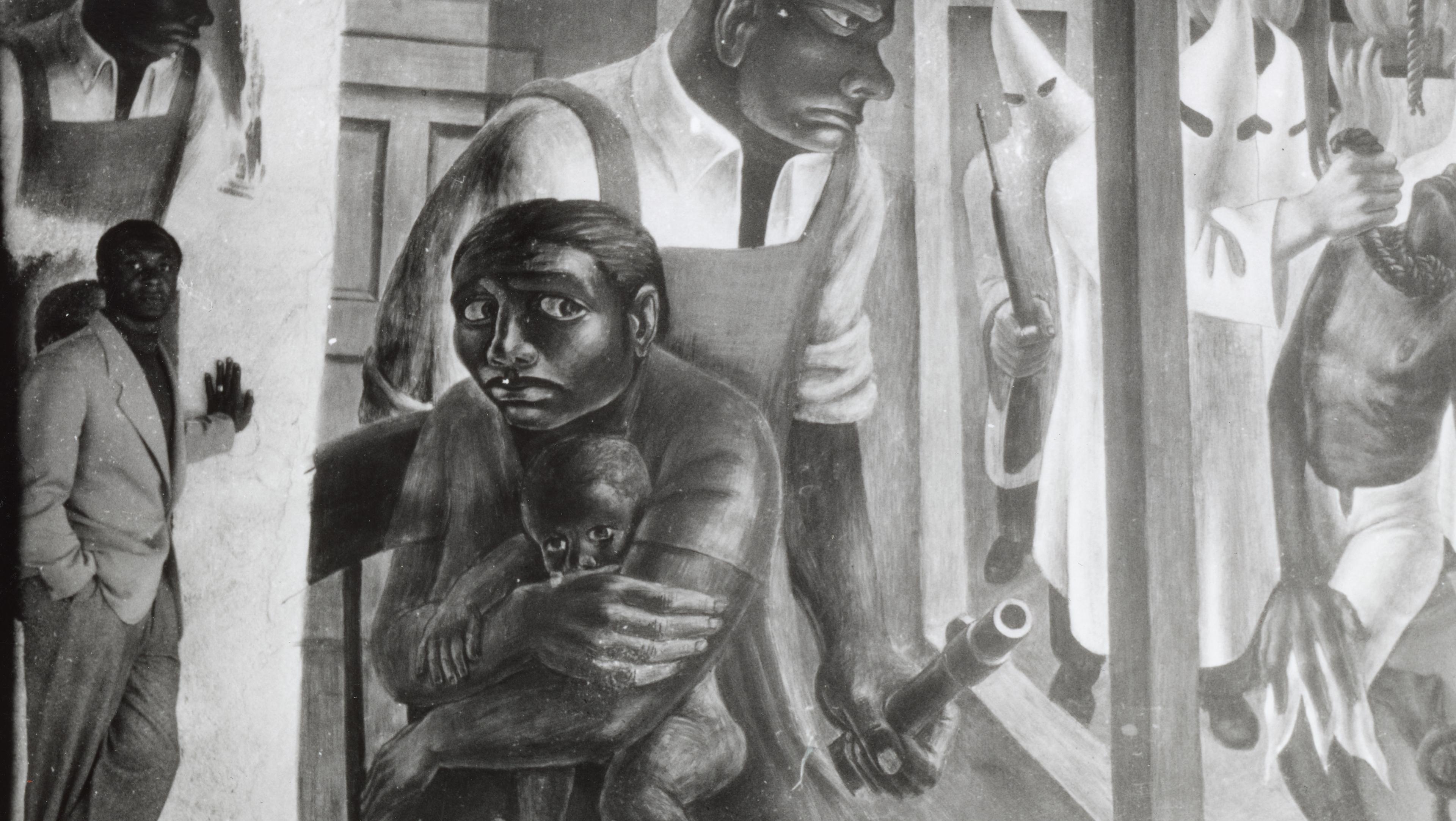 Black and white image of man standing next to mural depicted in black and white of a mother shielding her baby and while a strong black meale guards her. 