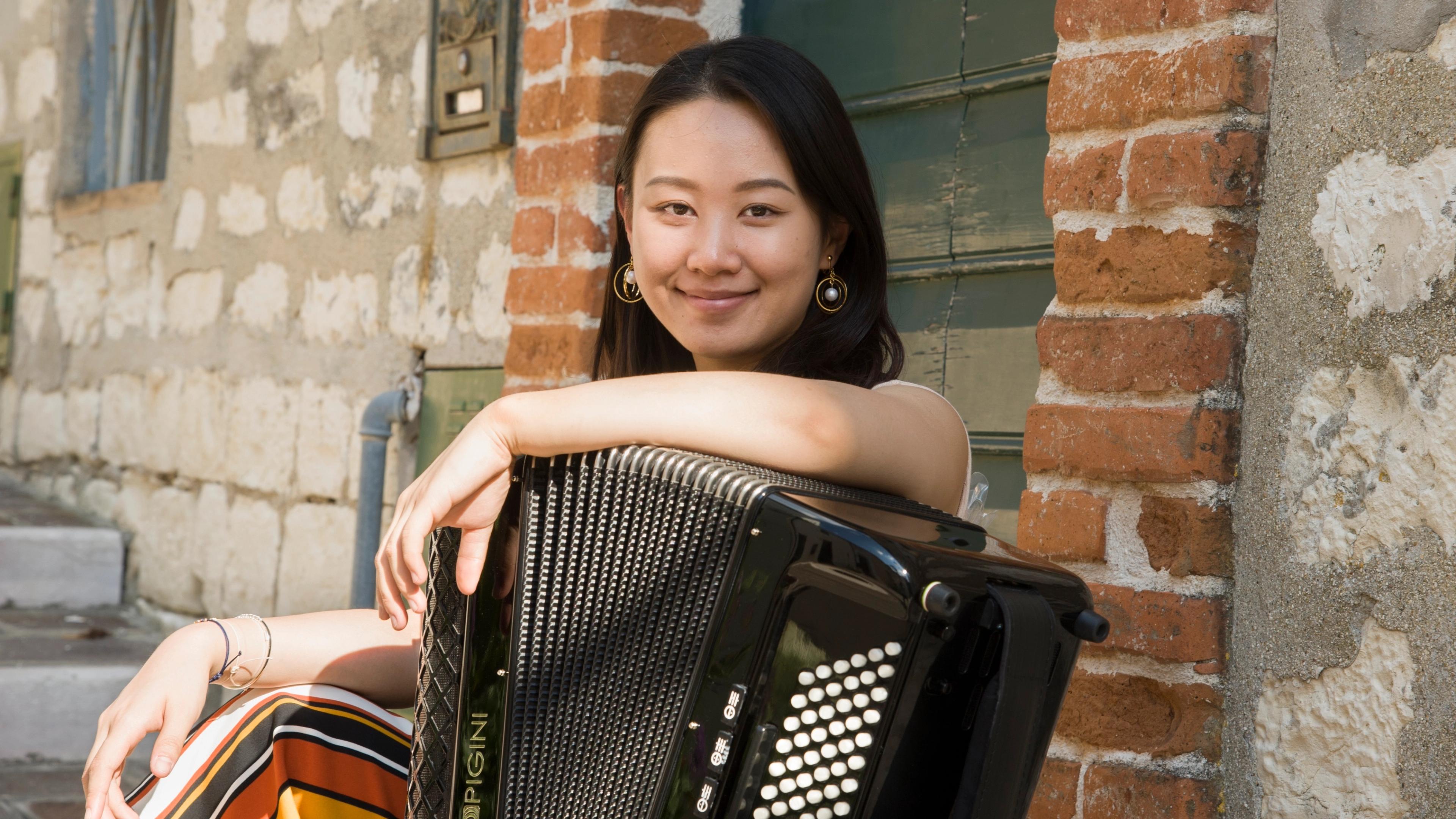 A woman smiles at the camera while holding an accordion. She sits against an exposed-brick backdrop.