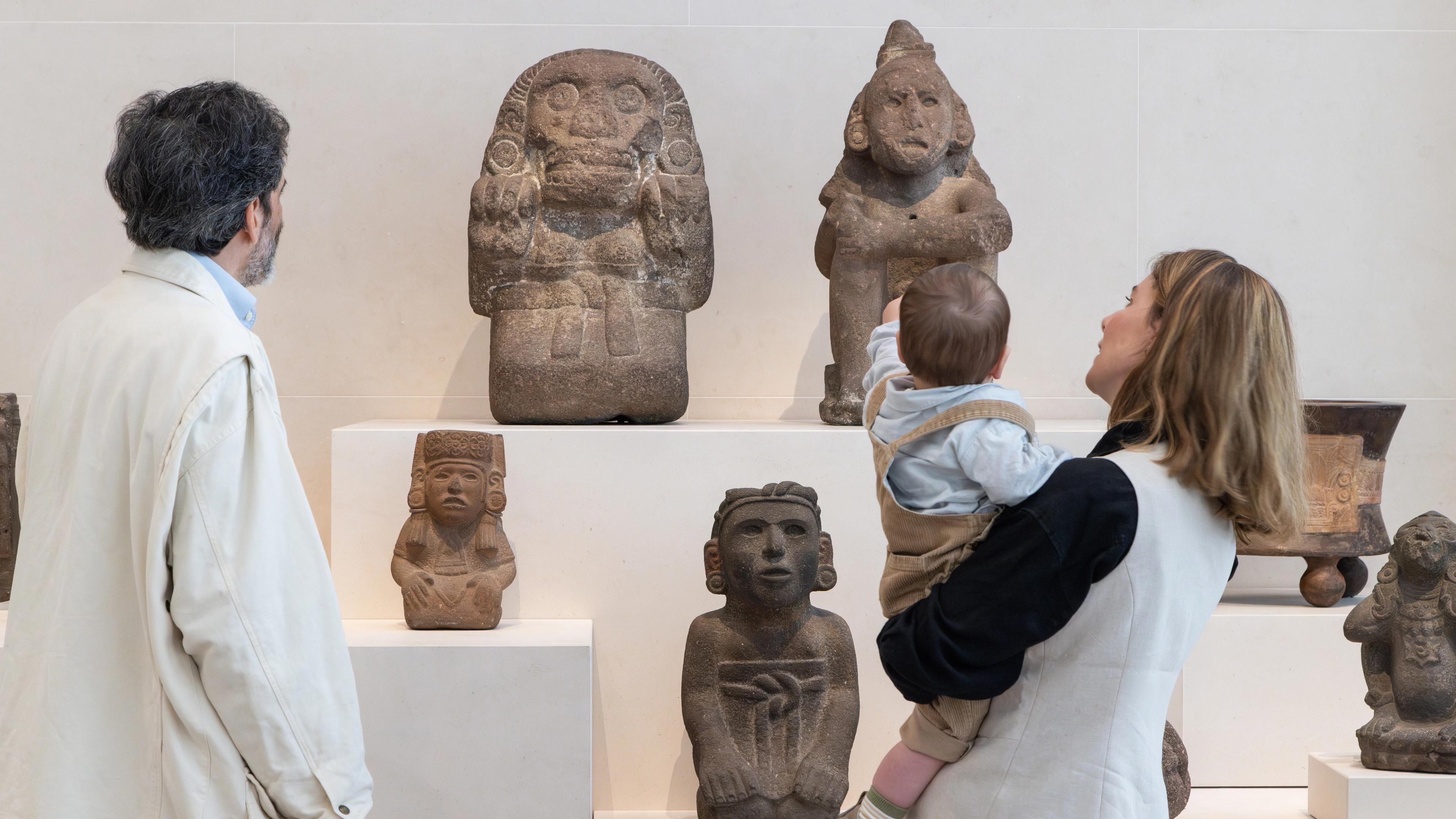 A man and a woman holding a toddler, who points at Mexica sculptures in Gallery 360, Arts of the Ancient Americas, The Michael C. Rockefeller Wing.