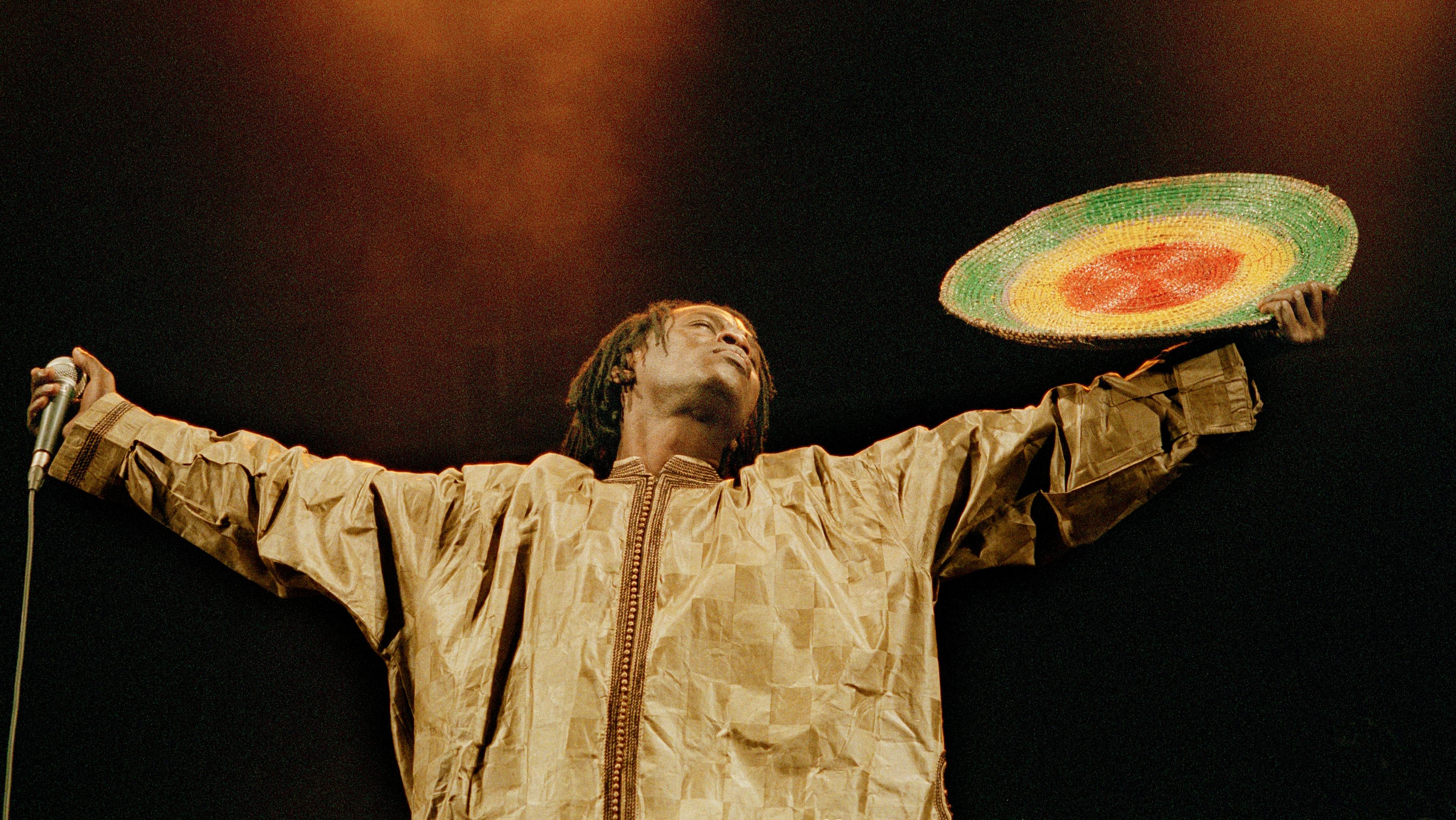 A man in a zip-up jacket holds his arms outstretched, facing his closed eyes toward the sky. He has a microphone in his right hand and a woven shield in his left.