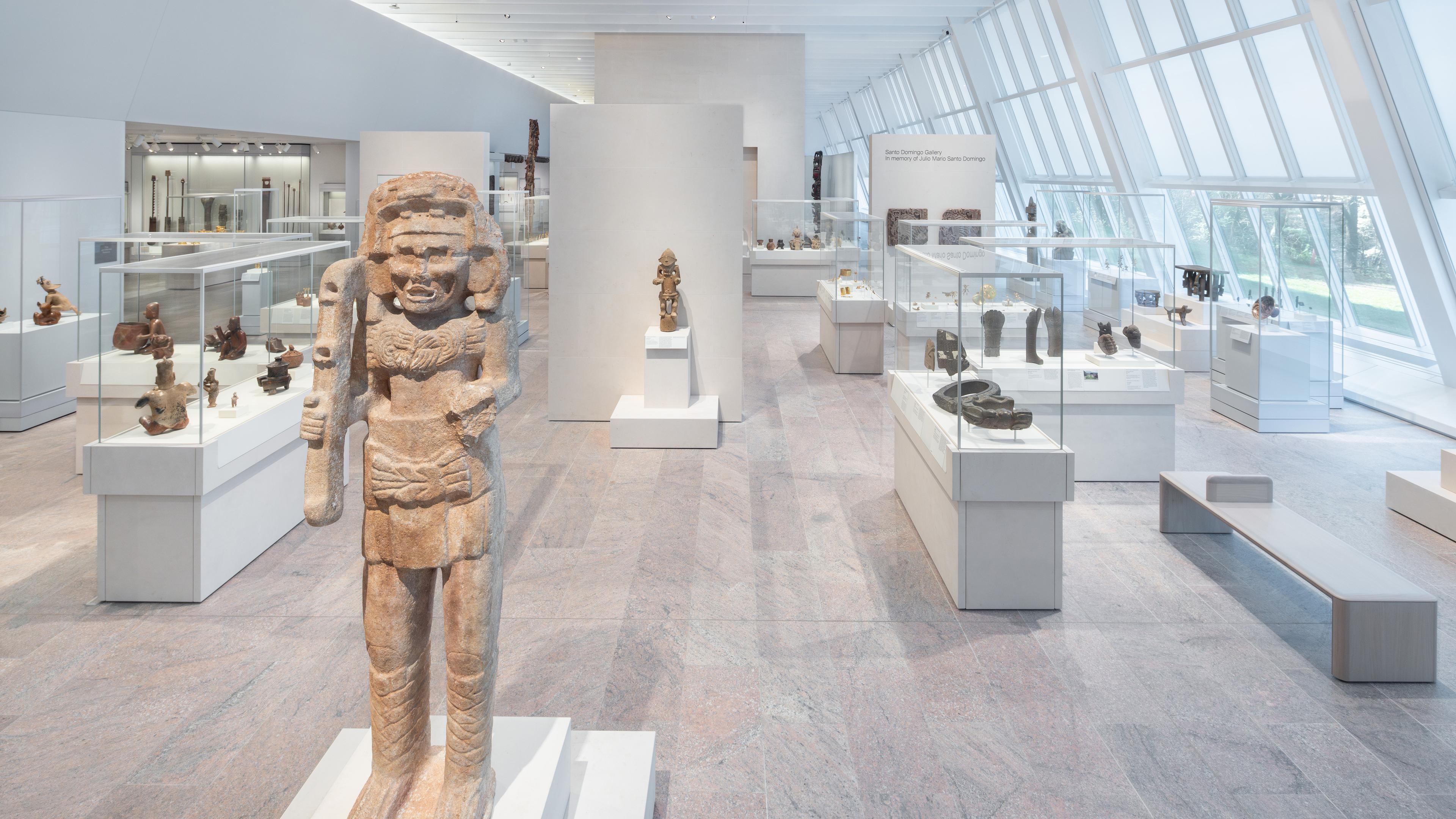 Bright gallery with white pedestals showcasing a variety of stone statues and objects in glass cases.