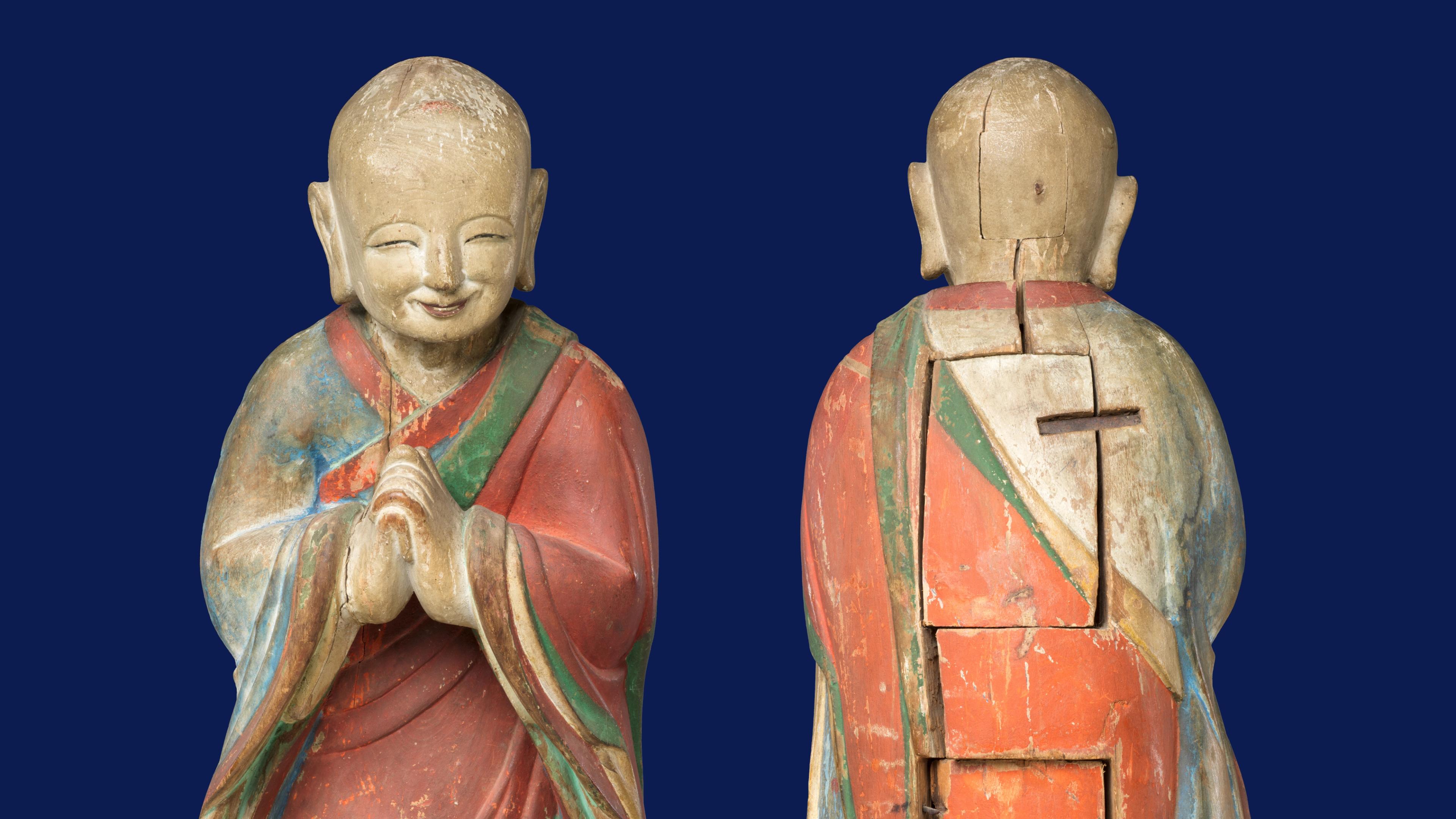 Carved wooden statue of a smiling monk in a red and blue robe, hands together in prayer. Front and back view on a dark blue background.