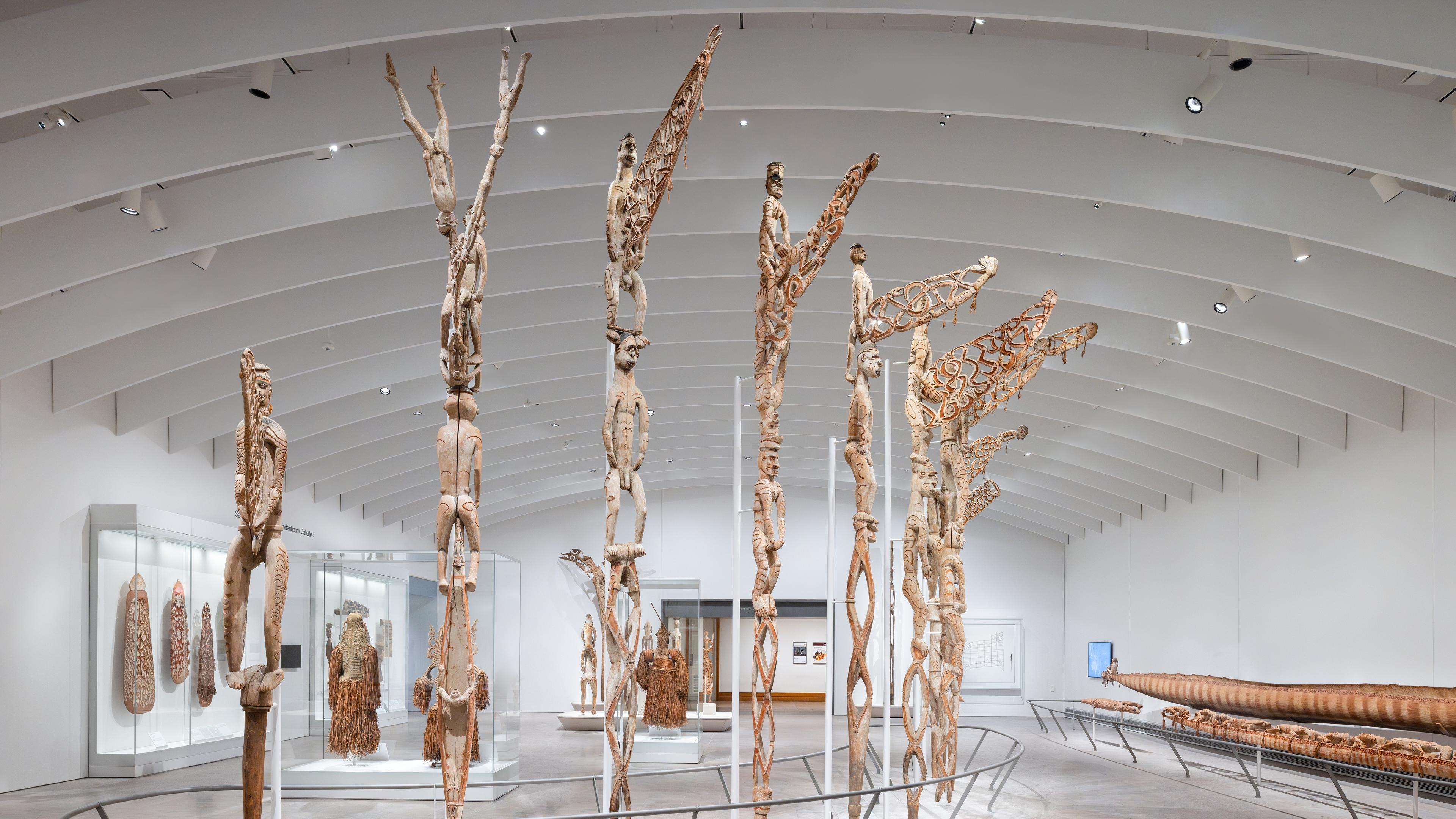Tall ancestor poles arranged in a curve in the center of a bright white gallery space.
