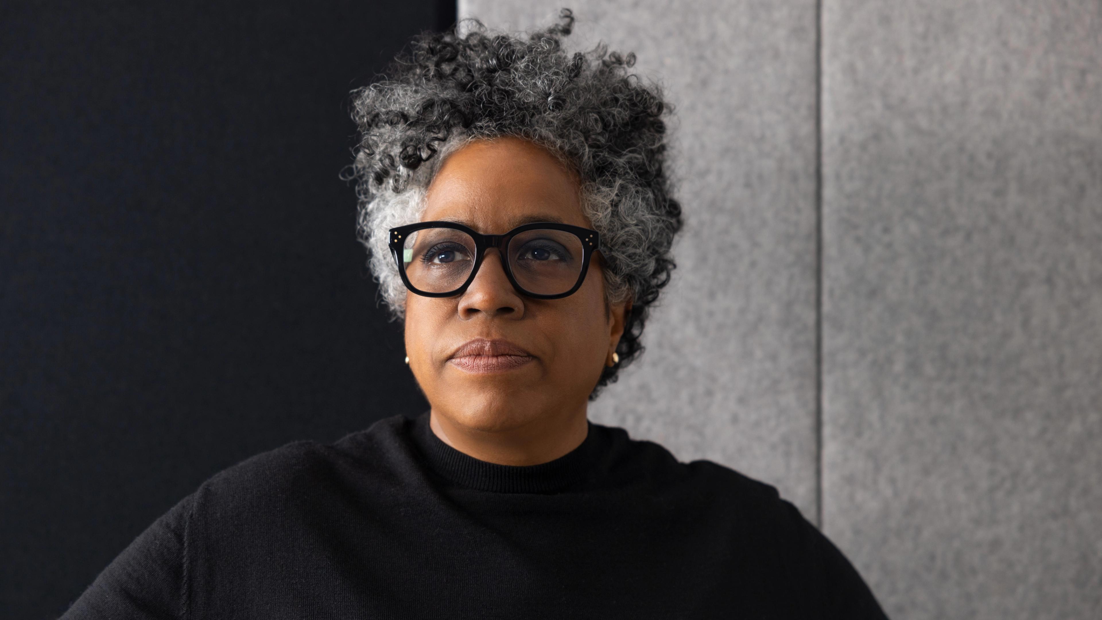 A woman wearing a black shirt and black glasses stares into the distance.