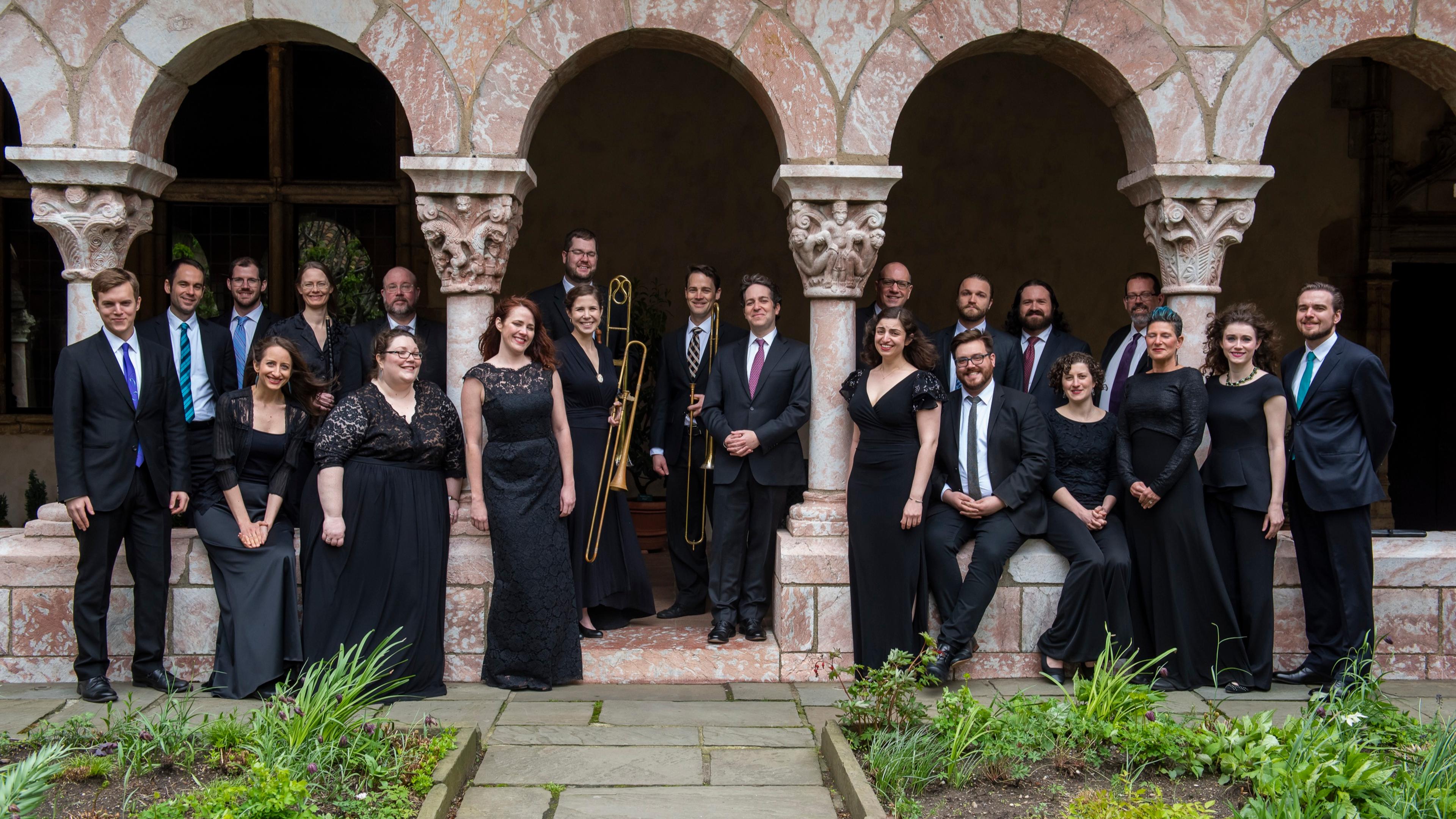 A choral ensemble poses for a photo in front of medieval archways.
