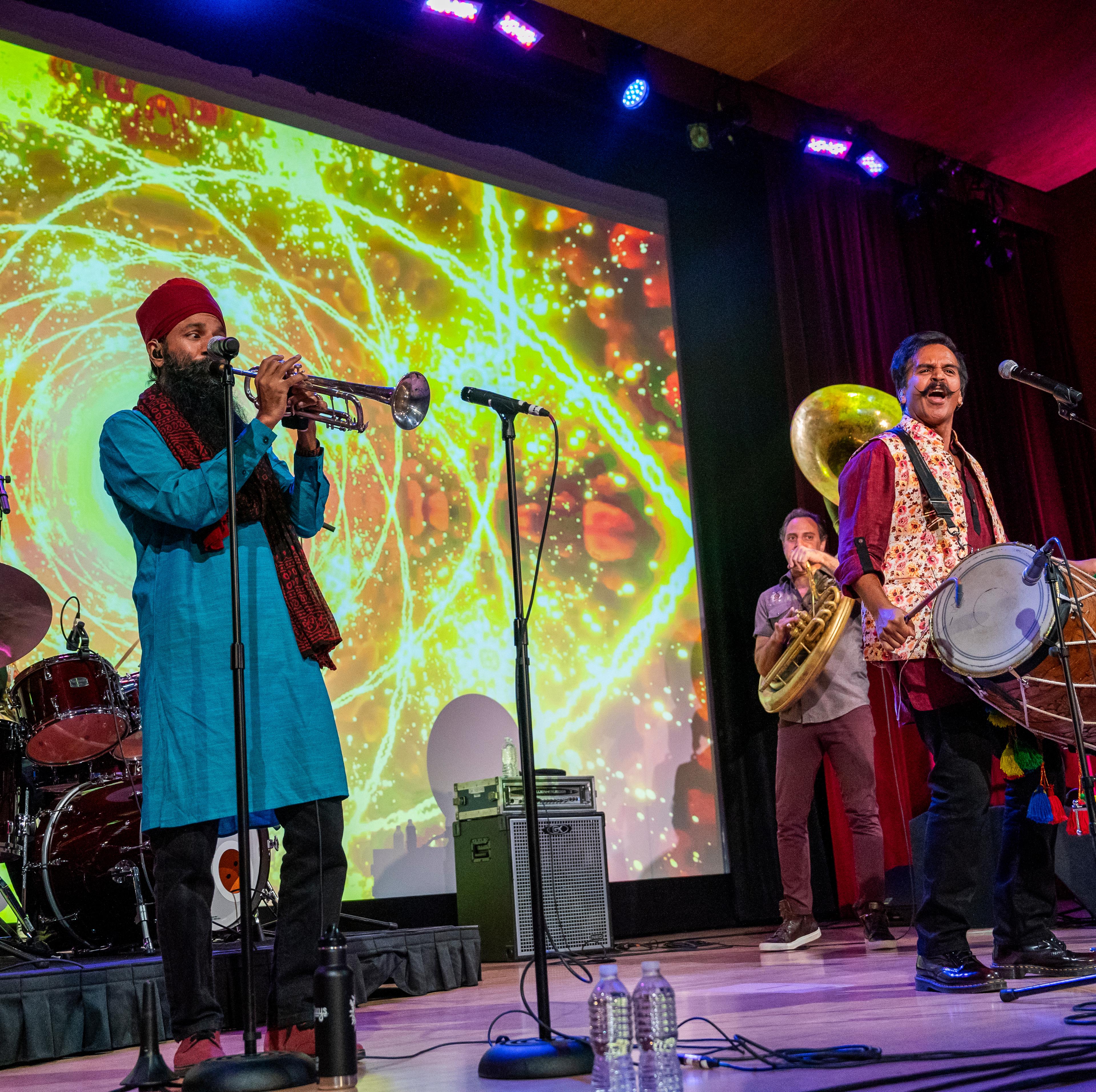 A drummer, a trumpet player, a dhol player, a sousaphone player, and a trombone player perform on a stage in front of psychedelic projections.