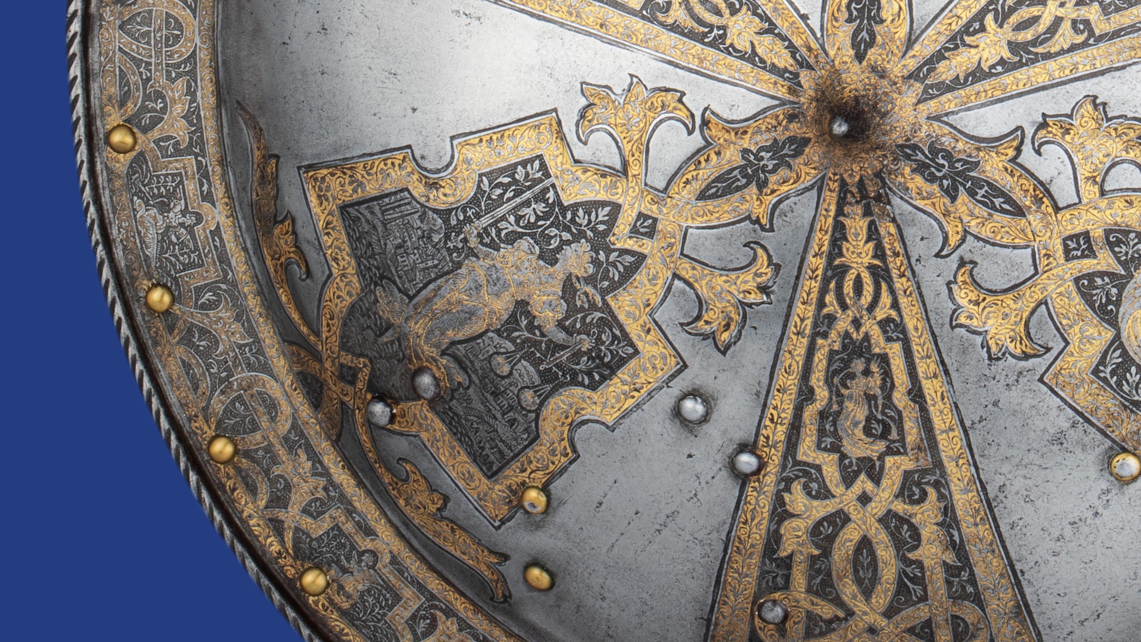 Ornate silver shield with intricate gold vine patterns and medieval animal motifs, accented by brass rivets on a blue background.