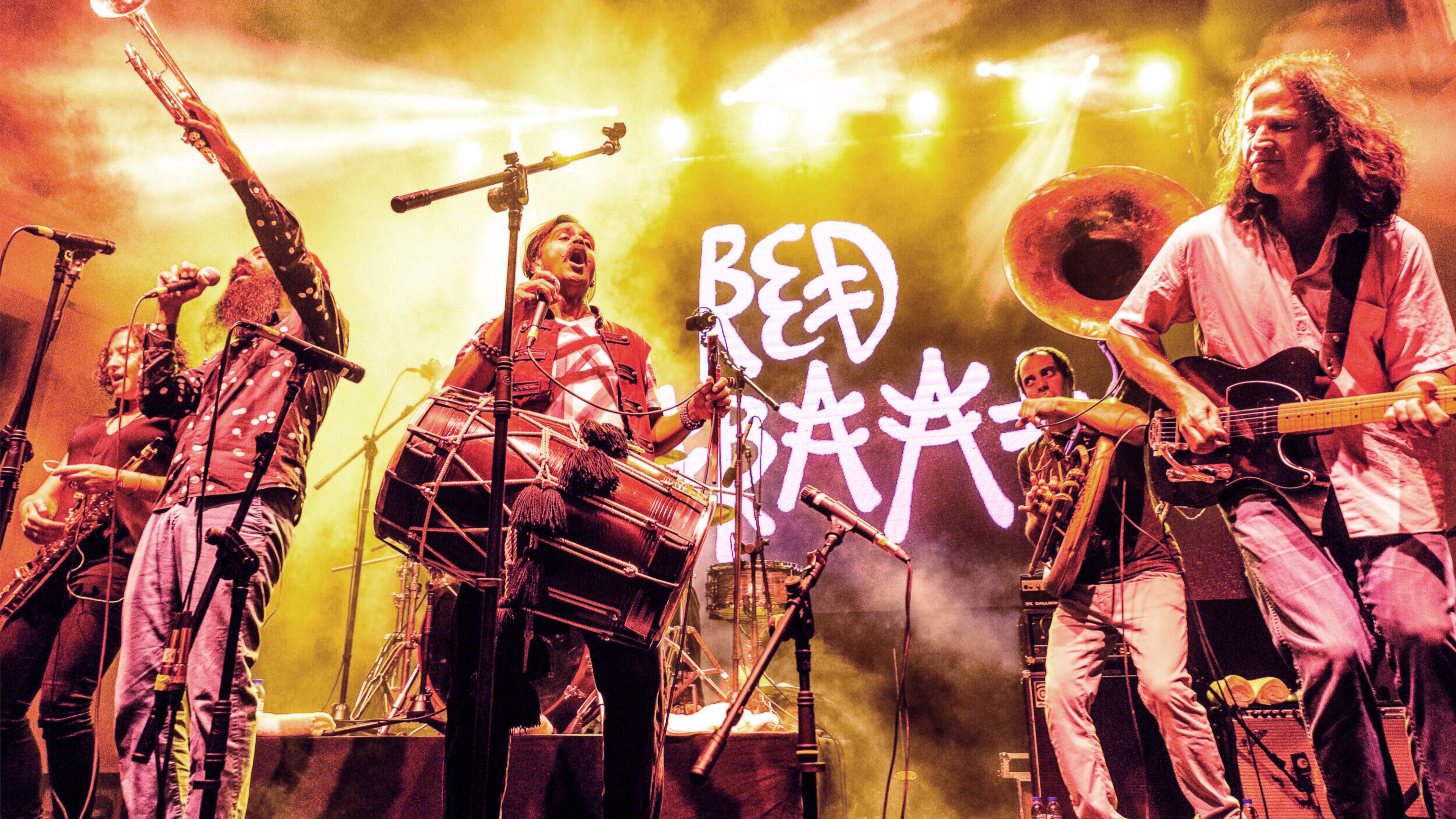 A band of brass players, led by a dhol player, rocks out in front of red light. The back wall reads "Red Baraat."